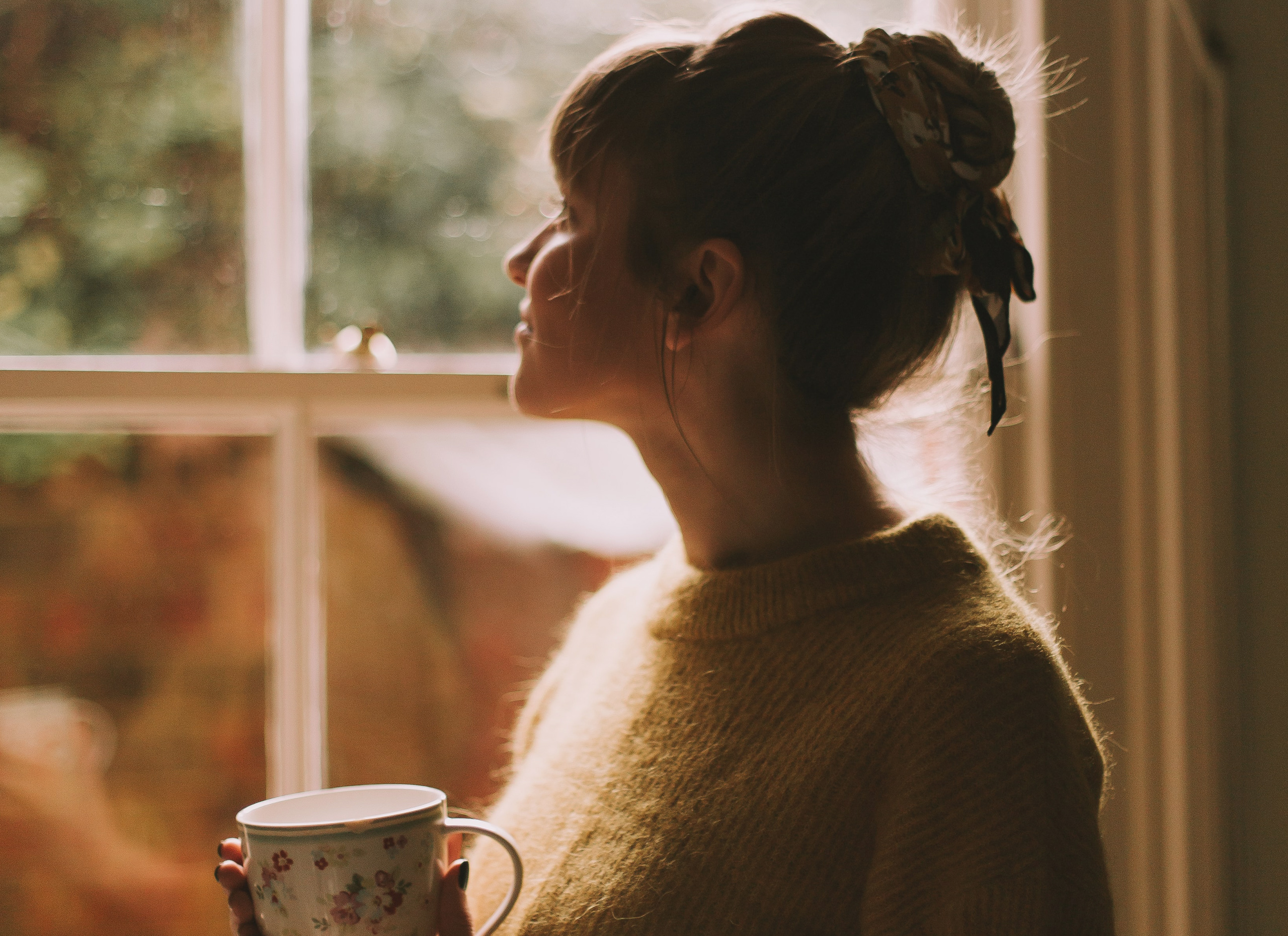 A woman is looking out a window and she is holding a mug in her hand.