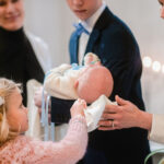 Pieni tyttö kuivaa papin kanssa kastetun vauvan päätä valkoisella liinalla.
