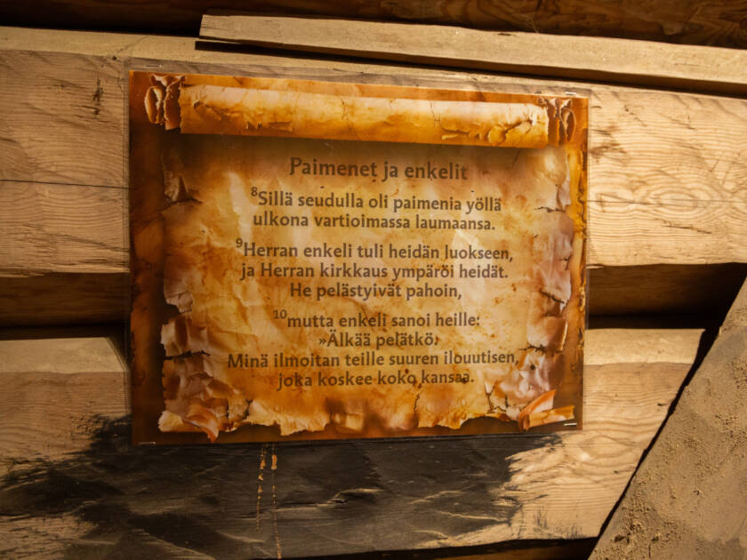 Jouluevankeliumiteksti kellotapulissa. Taustalla hirsiseinä.