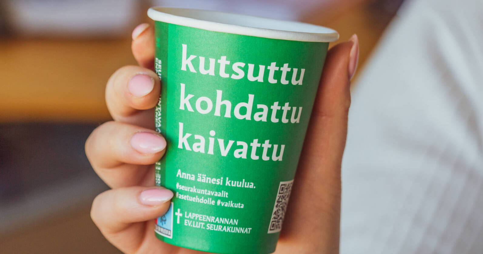 Nainen pitää kädessään vihreää pahvimukia, jossa sanat kutsuttu, kohdattu, kaivattu.