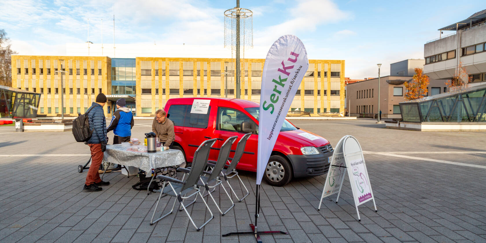 Serkku toiminta torilla. Taustalla punainen auto ja edessä kolme miestä sekä pisaran muotoin beach flag.